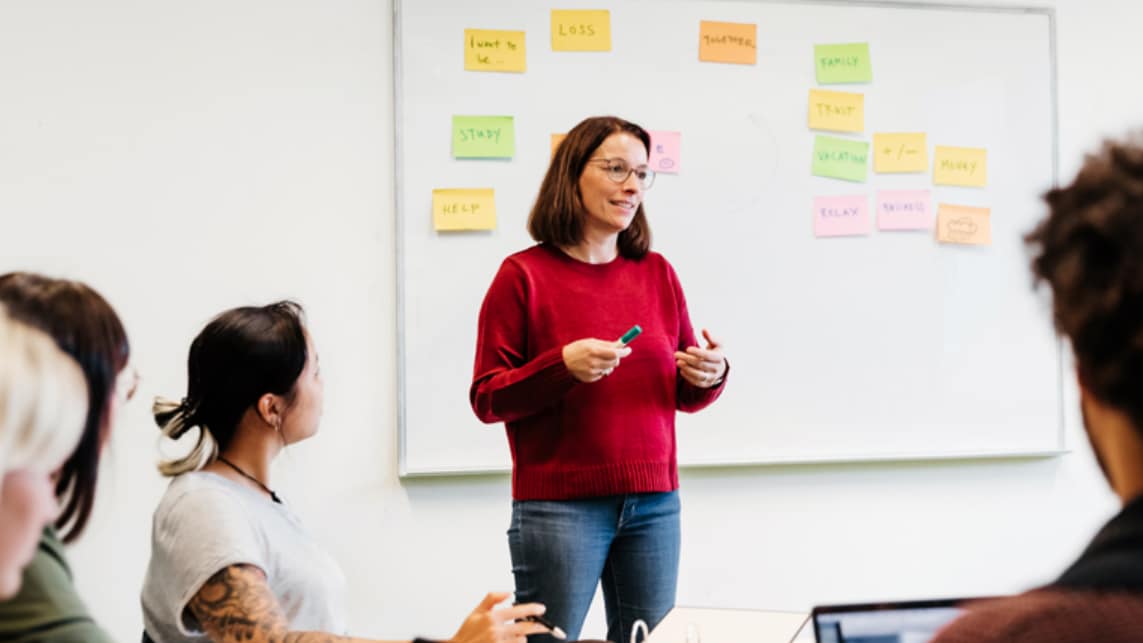 Woman discussing suicide prevention with group of people. Whiteboard with yellow, green, and pink note cards behind her.