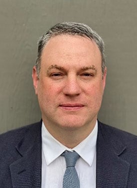 Headshot of Noah Burg wearing a dark colored suit with a solid neutral background.