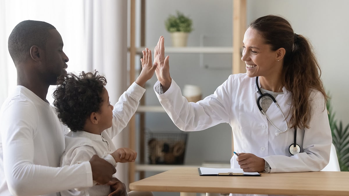 Female healthcare provider high fives young boy sitting on father's lap in doctor's office.