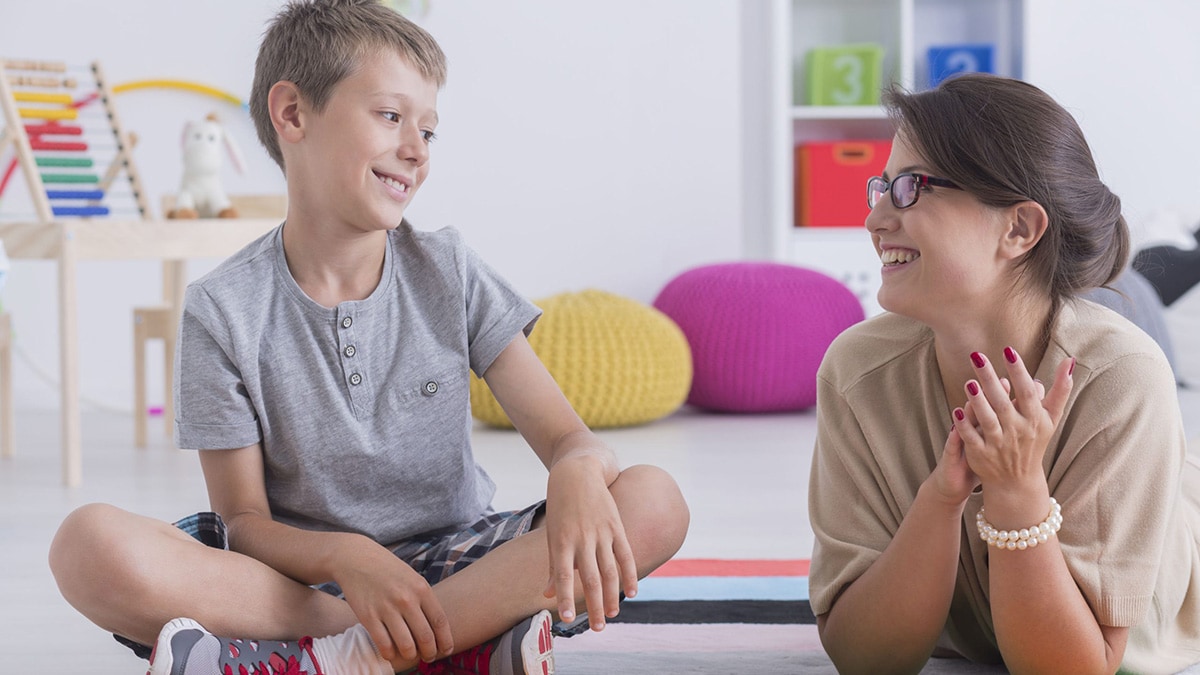 Una terapeuta acostada con los codos en el piso al lado de un niño sentado.