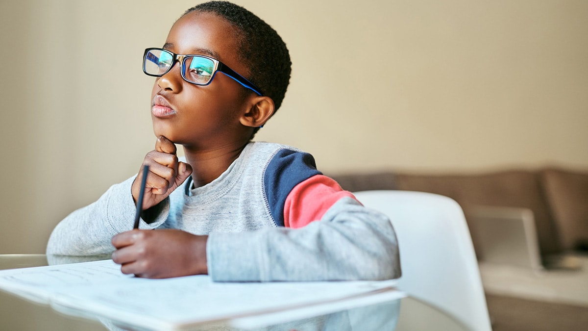 Boy sits at desk writing with his chin resting on his hand in a distracted way..