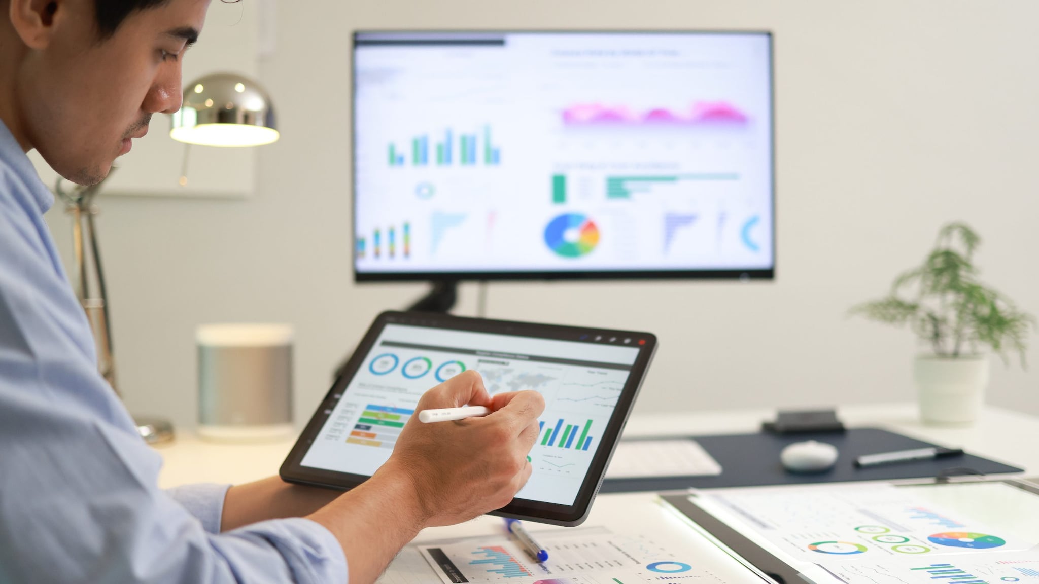 Health economist reviewing data and graphs on a computer monitor and tablet.