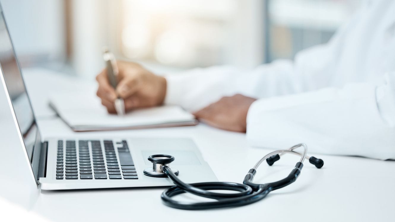 Desk with doctor writing on a pad with a computer and stethoscope in the foreground.