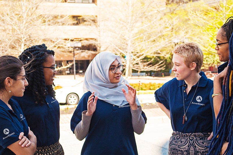 A diverse group of five female Public Health AmeriCorps members are outdoors having a conversation.