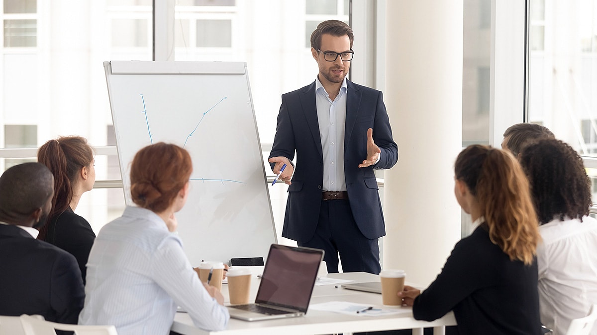 Man in suit standing in front of easel with hand-drawn chart in conference room full of employees.