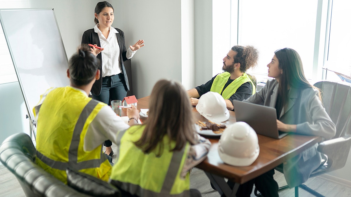 Businesswoman standing at dry erase board training three workers in hardhat gear and one woman in business attire.